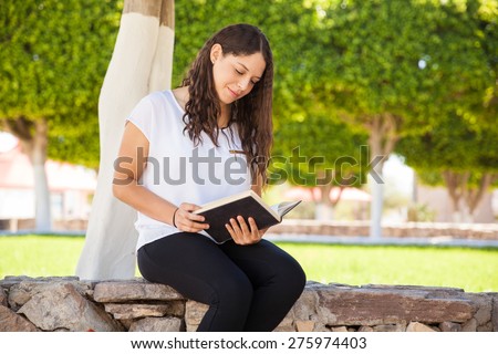 Cute brunette with curly hair reading a book while sitting outdoors at school