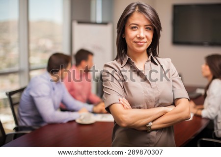 Portrait of a pretty young brunette dressed casual and standing in a meeting room with her co-workers in the background
