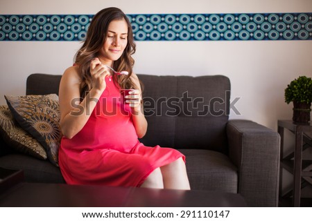 Pretty young pregnant woman eating some yogurt in the living room