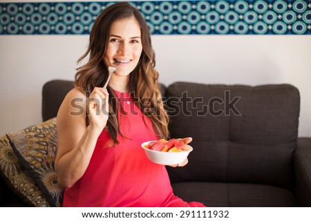 Cute young pregnant woman biting a fork while eating from a bowl of fruit at home