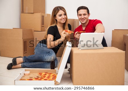 Cute young couple eating pizza and watching a TV show on a tablet computer while moving into their new home