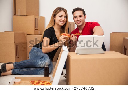 Portrait of a good looking Hispanic couple eating pizza and watching a show on a tablet computer while moving into a new apartment
