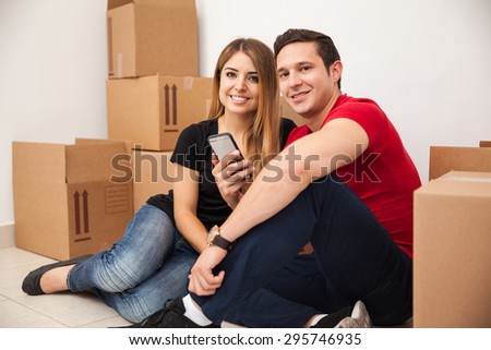 Good looking Hispanic married couple moving to their new home and taking a break to do some social networking on a phone