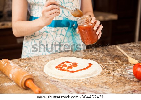 Closeup of a woman wearing an apron adding tomato sauce to her homemade pizza in the kitchen