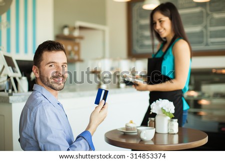 Handsome young Hispanic man holding a credit card after paying for his coffee and cupcake at a cafe and smiling