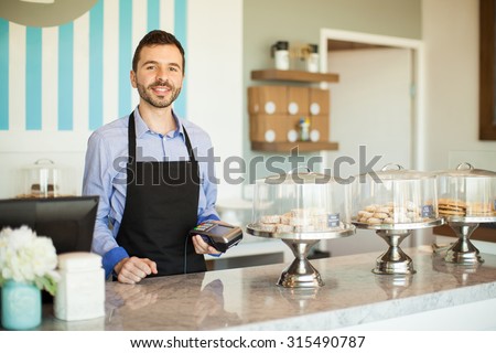 Attractive young Latin man holding a bank terminal next to a cash register in a bakery