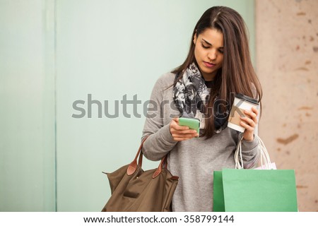 Beautiful young woman using her smartphone and drinking coffee while doing some shopping in a mall