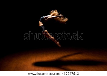 Full length profile view of a gorgeous female jazz dancer jumping during a performance in a dark stage under a spotlight
