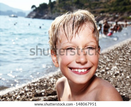Happy 8 year old blond boy in a swimming suit sunbathes on the beach and laughs. Sea in the background.