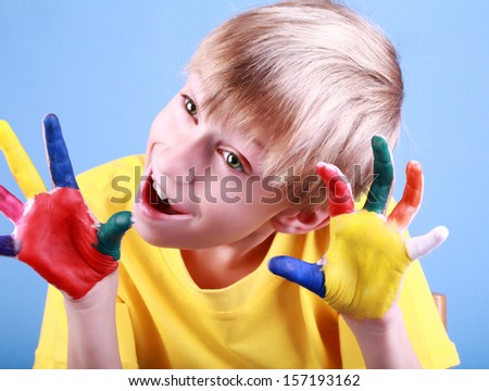 Beautiful cheerful blond boy in a yellow t-shirt raising his painted hands above his head and smiling happily over blue background