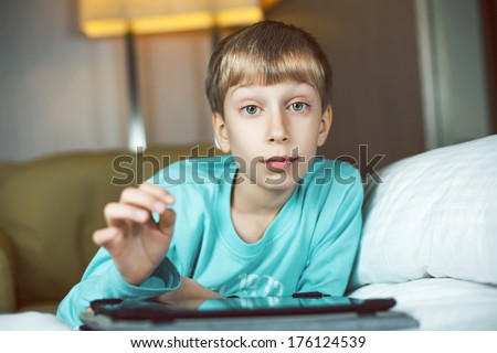 Beautiful child comfortably lying on a cozy bed playing at tablet computer looking into the camera