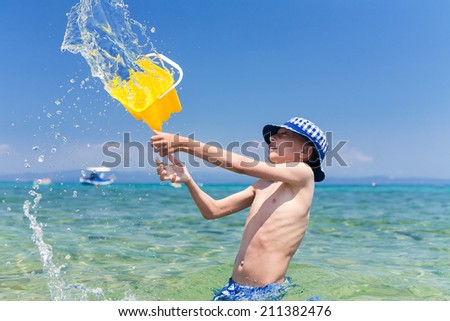 Funny little boy with a panama throwing a yellow toy bucket of water in the sea.