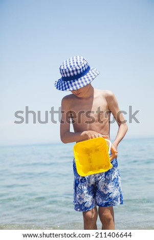 Child in swimmer shorts standing on beach holding a toy bucket pouring water