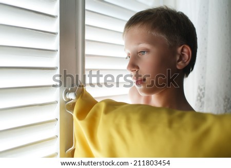 Candid portrait of a beautiful little child with a pillow standing by a window with blinds looking sad.