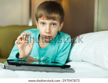 Cute funny little child playing at tablet computer sitting on cozy bed looking into camera.