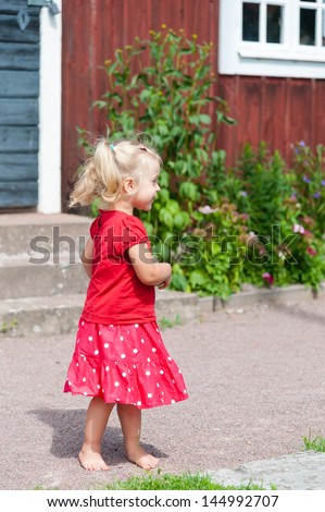 Little blond girl with pigtails in a red summer dress in the garden in front of a typical red wooden house in Sweden