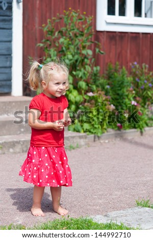Little blond girl with pigtails in a red summer dress in the garden in front of a typical red wooden house in Sweden
