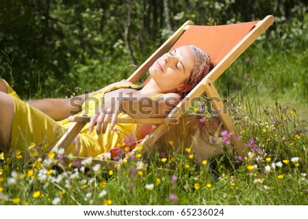 A young woman relaxing in a deck chair. A cat under the deck chair is resting too. The whole scene took place on an idyllic summer meadow in the swedish countryside