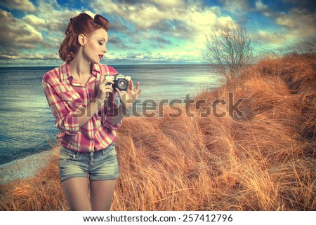 pin-up with a camera. Sexy redhead woman near the sea despite the weather photographs on camera nature. vintage retro