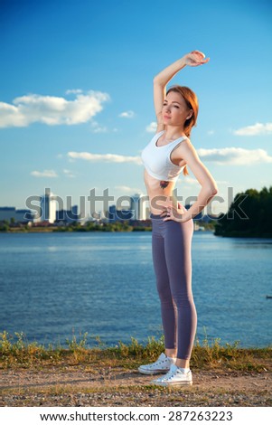 red-haired girl in the evening at sunset near the water on the background of the city makes the exercise in for sports and relaxation