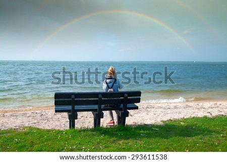 girl sitting on a bench near the sea and looking at the rainbow in the sky