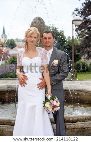 beautiful young wedding couple, blonde bride with flower and her groom against fountain