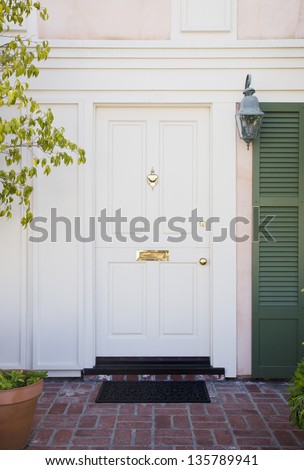Front door of an upscale home/Vertical shot of a white front door with brass accents and green stutters on a home with view of brick, mail slot and door mat.