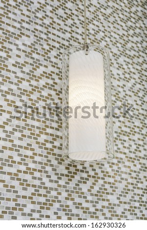 A lit light fixture hanging from the ceiling against a mosaic tile wall in a bathroom.