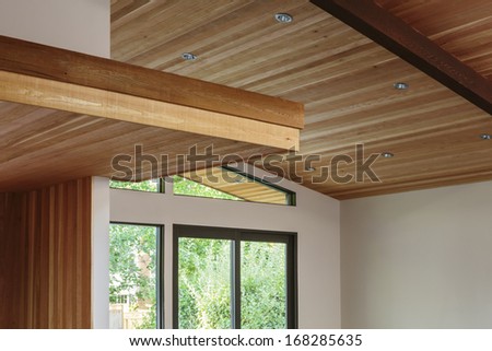 Detail of sloped wood beam ceiling with supports and wooden platform in house entryway. Trees can be seen outside the windows.