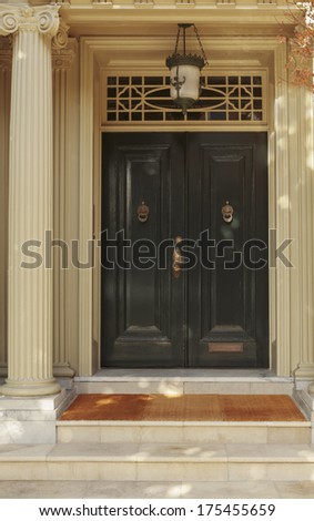Closed ornate wood door of an upscale home, accented with an iron barred window, iron door knocker, and iron bolts. The door is set into a beige stucco house, framed with a wide arch and planters.