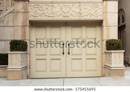 Closed ornate wood door of an upscale home, accented with an iron barred window, iron door knocker, and iron bolts. The door is set into a beige stucco house, framed with a wide arch and planters.