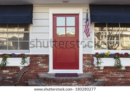 A red front door of an American home. Also seen is white siding, American flag, blue awning, flowers, and brick steps.