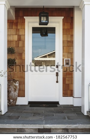 A white front door of a luxury home. The home is naturally stained wood shingles. The door is flanked by two white columns. Also seen is a stone porch and steps.
