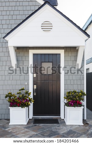 Glossy front door to a family home; The door is made of vertical wood boards, with a window. It is framed by two flower planters, gray shingles of the house, and a door mat.