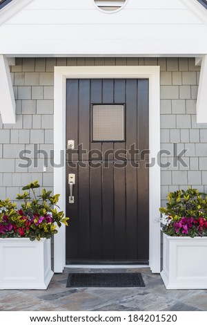 Glossy front door to a family home; The door is made of vertical wood boards, with a window. It is framed by two flower planters, gray shingles of the house, and a door mat.