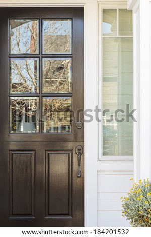Crop of a black, lacquered front door to a white family home, with white detail. The door has six windows. Also seen is a plant with yellow flowers.