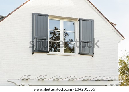 An upwards view of a white family home\'s top-floor window, with green shutters. The house is white brick. Some shingles, a reflection of a tree, and a clear sky can also be seen.