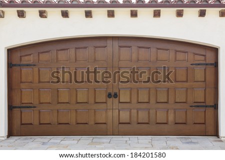 Double wooden swinging doors to a family home garage. The home is peach stucco. Also seen is the stone driveway and roof detail with shingles and wood beams.
