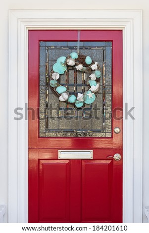 Detail of lacquered red front door with a white-and-blue seashell wreath. The door has an ornate, etched glass pane, and a mailbox.