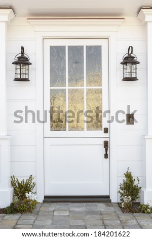 Detail of a white front door to a classic white family home. Door features intricate etched detail. Also seen is columns, a stone porch, and light fixtures.
