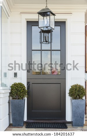A black front door to a white family home in daytime with door mat, light fixture, plants in blue planters, and a hint of a large window.
