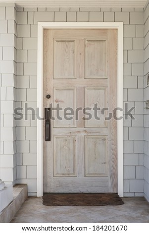 Detail of a wooden front door to a family home with raw, natural finish. House is white with shingles. Also seen is doormat, white column, and stone tile porch.