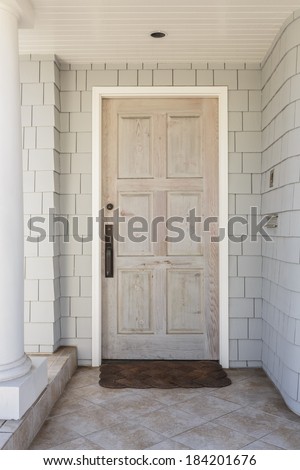 A front porch to a white home, with a wooden front door. Door has a raw, natural finish. House has shingles. Also seen is doormat, white column, and stone tile porch.