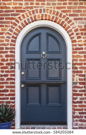 Glossy blue door to classic brownstone home with plant. A lacquered blue front door to a classic brownstone family home. Door is featured under brick arch, with door knocker, and a plant in blue pot.