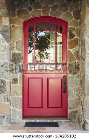 Detail of a red front door to a home in daytime. The door is arched with glass windows, and is framed by the house\'s stone detail. Also seen is greenery, a doormat, and the reflection of a tree.