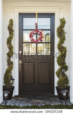 Black front door of home with heart wreath. Black front door to a home in daytime. Door is framed by two plants, and adorned with a wreath of pink and red hearts.