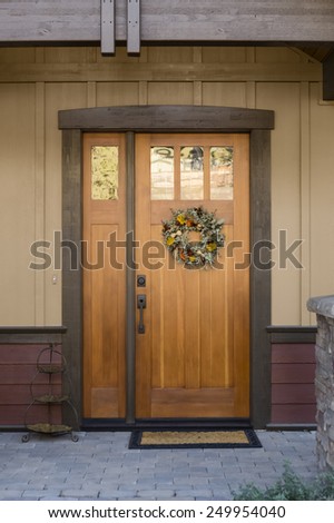 Warm Entryway with Natural Wood Front Door, Hanging Wreath, Doormat and Decorative Piece on Building with Tan Paneled and Red Siding Exterior