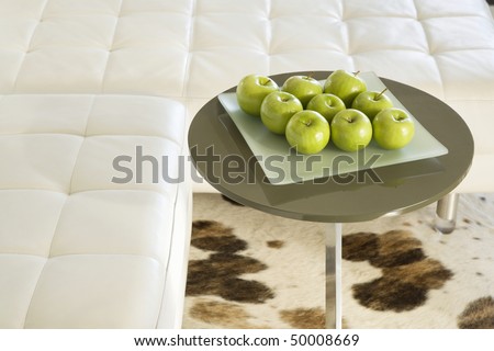 Green apples sitting on a square frosted glass plate on a round table. The table is standing on a spotted rug and is next to a white sectional sofa. Horizontal shot.