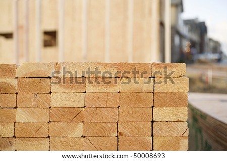 Selective focus view of the end of a stack of 2x4 boards at a construction site, with a framed building and finished construction in the background. Horizontal shot.