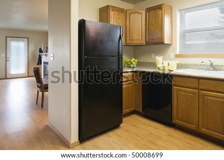 View of a kitchen with a black refrigerator and wood cabinets, showing a partial view of the dining room and living room areas. Horizontal shot.
